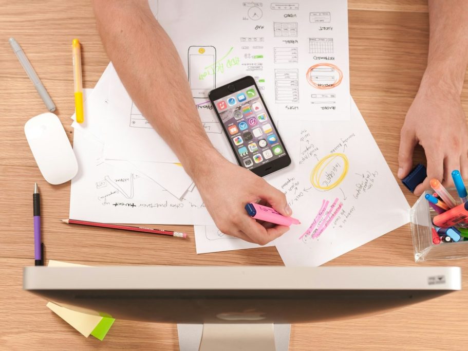 Hände, die an einem Schreibtisch mit Plänen, einem Smartphone und Stiften arbeiten.